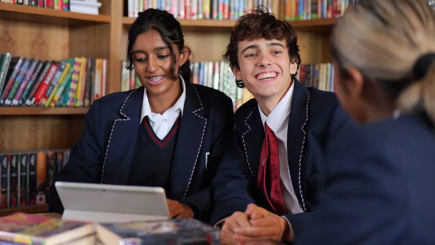 Two pupils in school uniform smiling over a tablet in the library